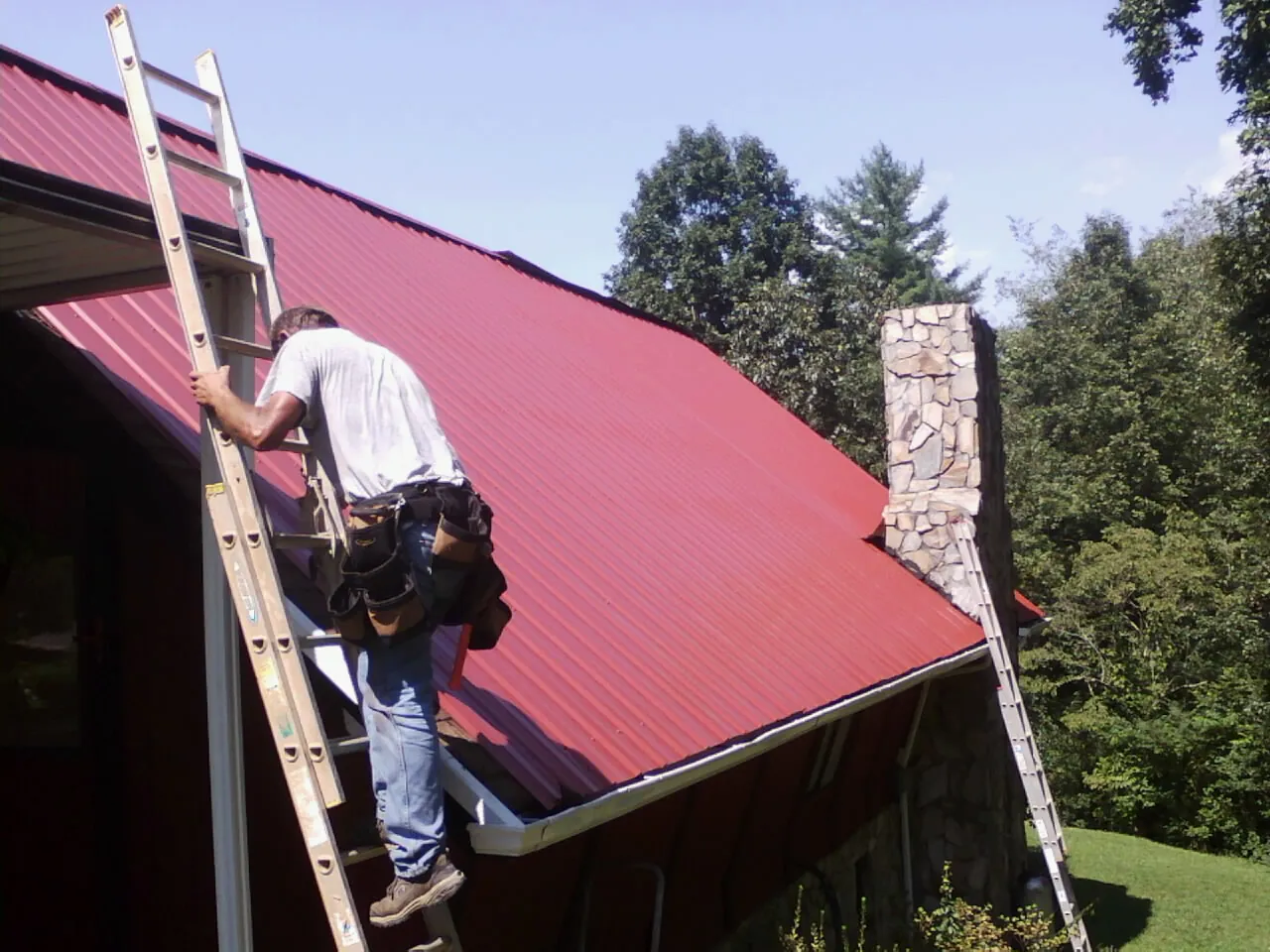 Roofer on a ladder beside a red metal roof and stone chimney — Soffit & Fascia Repair by Roofing Brothers in Palos Park